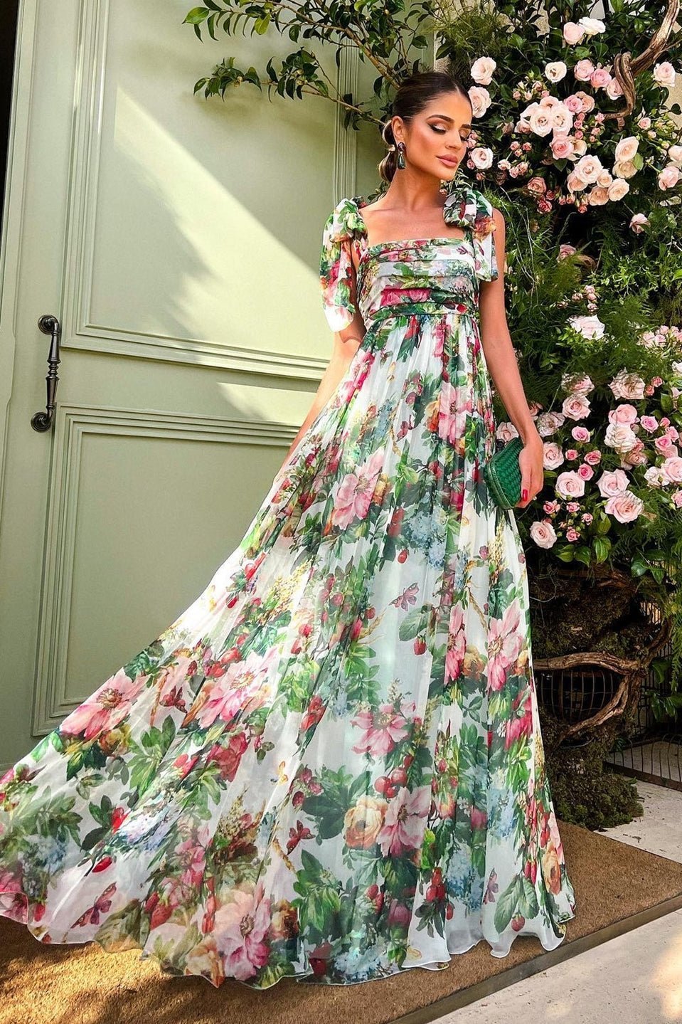 Multi - A woman models a flowing maxi dress featuring a vibrant, large-scale botanical floral print, styled with a ruched bodice and wide tie-shoulder straps, standing outdoors near a floral arrangement.