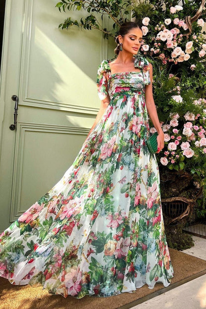 Multi - A woman models a flowing maxi dress featuring a vibrant, large-scale botanical floral print, styled with a ruched bodice and wide tie-shoulder straps, standing outdoors near a floral arrangement.