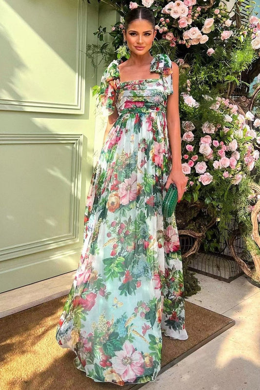 Multi - A woman models a flowing maxi dress featuring a vibrant, large-scale botanical floral print, styled with a ruched bodice and wide tie-shoulder straps, standing outdoors near a floral arrangement.