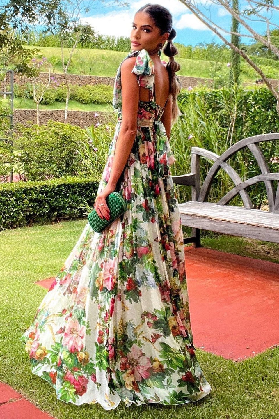 Multi - A woman models a flowing maxi dress featuring a vibrant, large-scale botanical floral print, styled with a ruched bodice and wide tie-shoulder straps, standing outdoors near a floral arrangement.