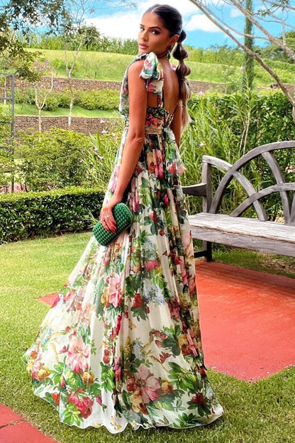 Multi - A woman models a flowing maxi dress featuring a vibrant, large-scale botanical floral print, styled with a ruched bodice and wide tie-shoulder straps, standing outdoors near a floral arrangement.