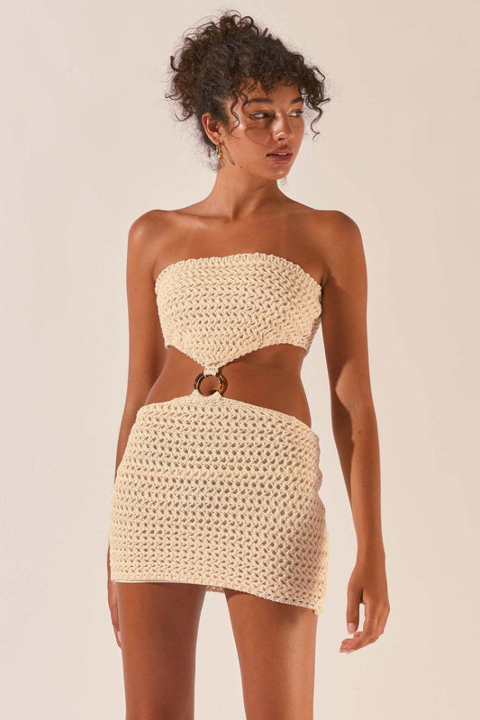 Beige - Front view of a model wearing a strapless, cream crochet mini dress featuring dramatic waist cut-outs connected by a central tortoiseshell-style ring against a plain studio background.
