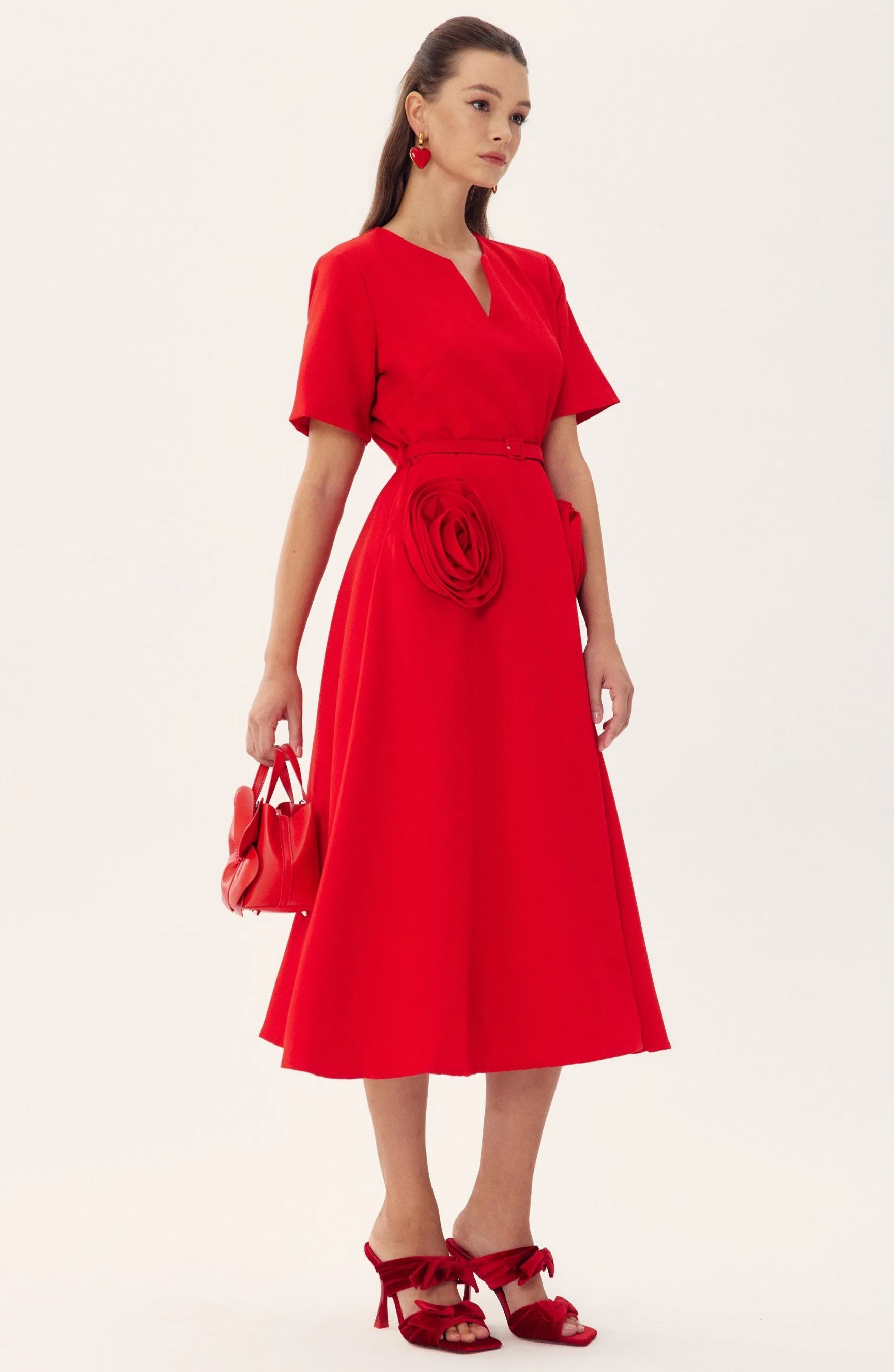 Red - A woman wearing a bright red, short-sleeved midi dress featuring a notched V-neckline, a thin waist belt, and two large 3D rosette embellishments on the A-line skirt.