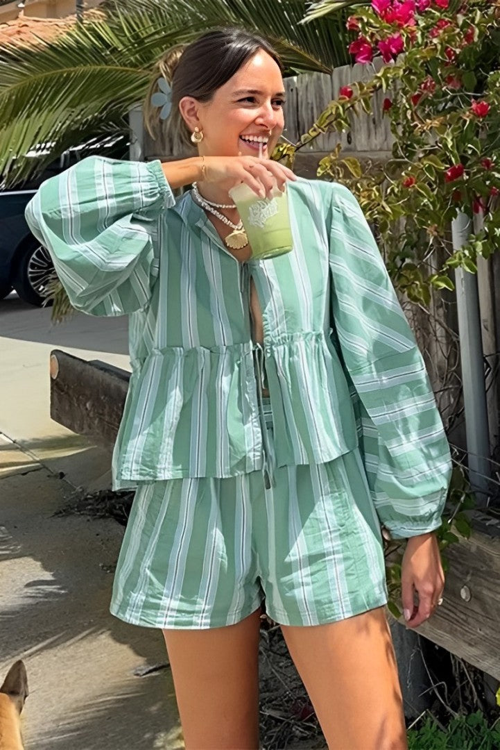 Green - A woman wearing a relaxed-fit, green and white vertically striped two-piece set featuring a tiered peplum top with long balloon sleeves and a tie-front closure, paired with matching loose shorts, standing outdoors.