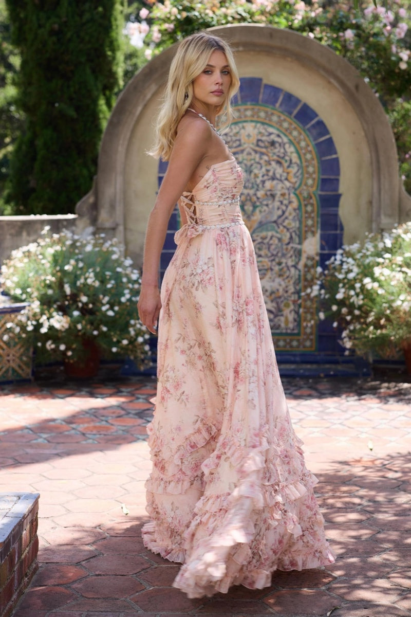 Pink - A woman stands outdoors in a garden setting wearing a light blue, floral print maxi dress featuring a beaded plunging halter neckline, a beaded waist, a high side slit, and a ruffled hem.