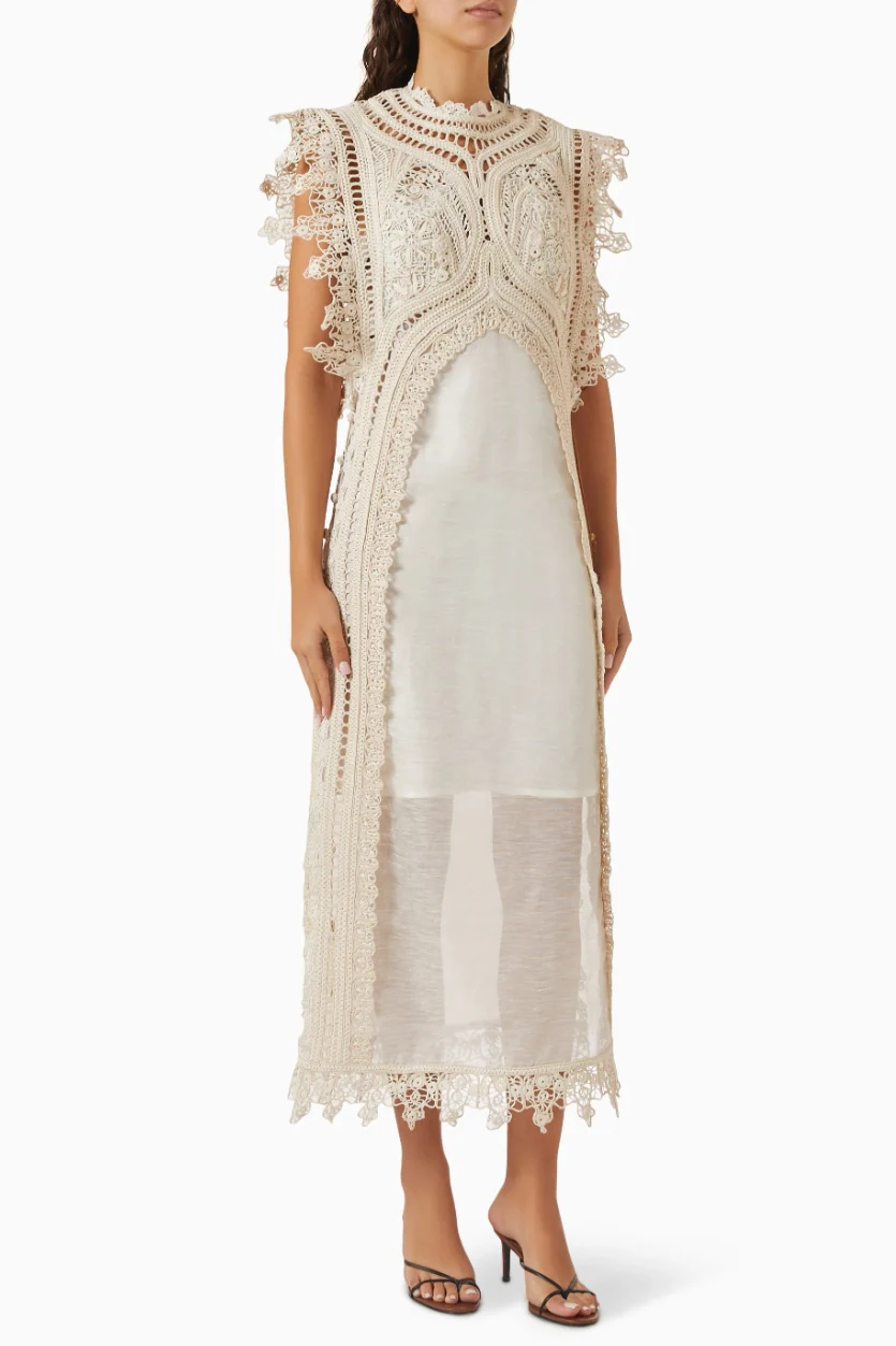 A woman wearing a cream-coloured midi dress featuring an intricate crochet overlay with scalloped edges over a semi-sheer slip.