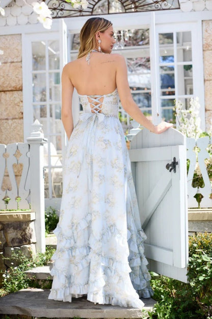 Blue - A woman stands outdoors in a garden setting wearing a light blue, floral print maxi dress featuring a beaded plunging halter neckline, a beaded waist, a high side slit, and a ruffled hem.