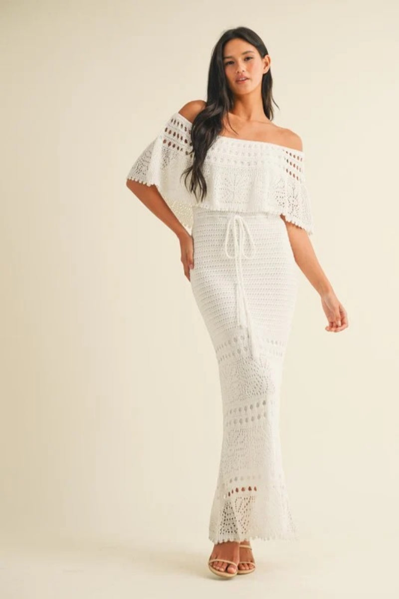 White - A woman wearing a white, off-the-shoulder crochet maxi dress with a ruffle neckline and tie-waist detail, standing against a neutral studio background.