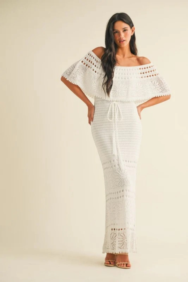 White - A woman wearing a white, off-the-shoulder crochet maxi dress with a ruffle neckline and tie-waist detail, standing against a neutral studio background.