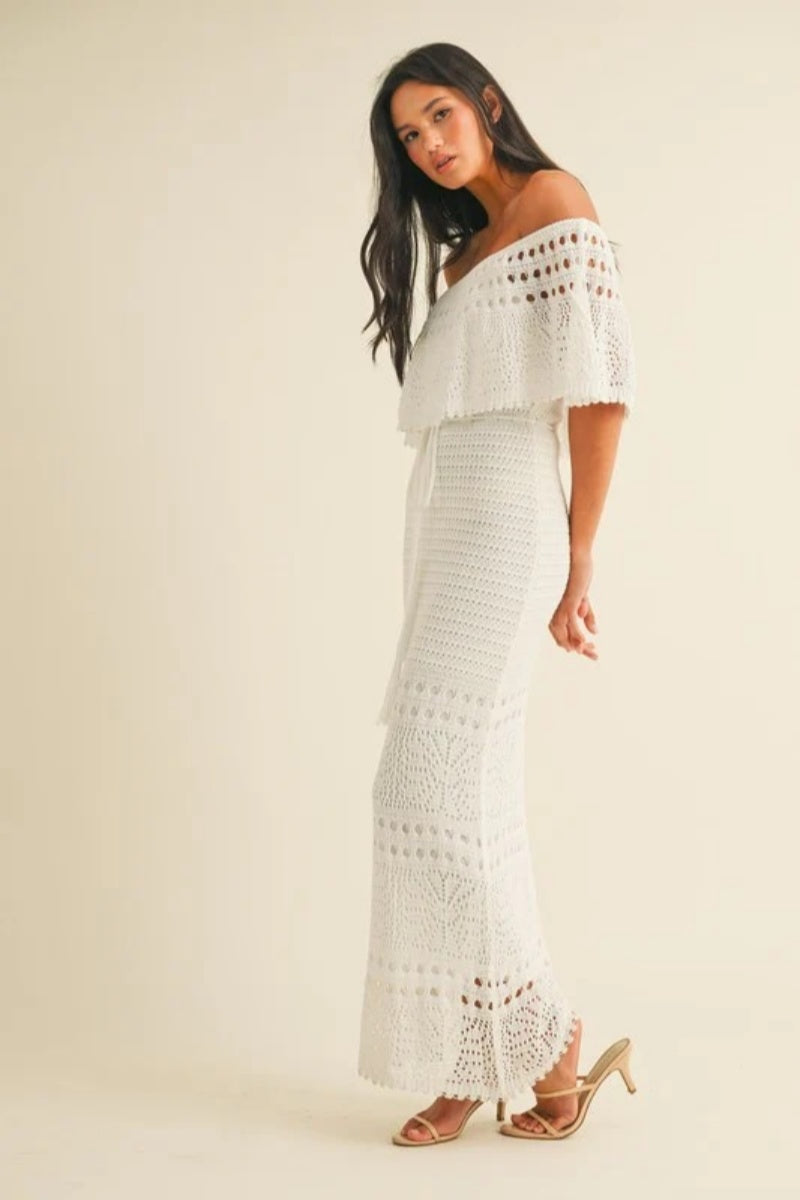 White - A woman wearing a white, off-the-shoulder crochet maxi dress with a ruffle neckline and tie-waist detail, standing against a neutral studio background.