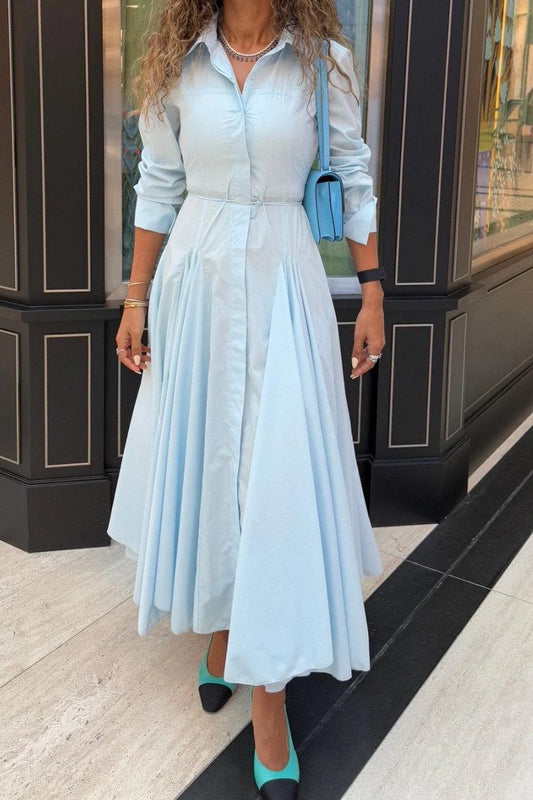 Lake Blue - A woman wearing a light blue, long-sleeved midi shirt dress featuring a collared neckline, button-down front, tie waist, and a voluminous pleated skirt with an asymmetric hem.
