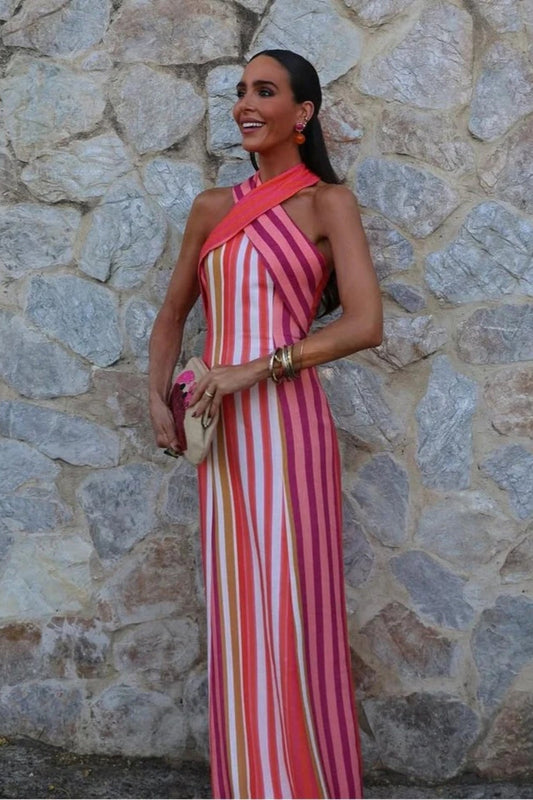 Red - A woman wearing a vibrant pink, orange, and white vertically striped maxi dress featuring a crossover halter neckline, posing outdoors against a textured stone wall.