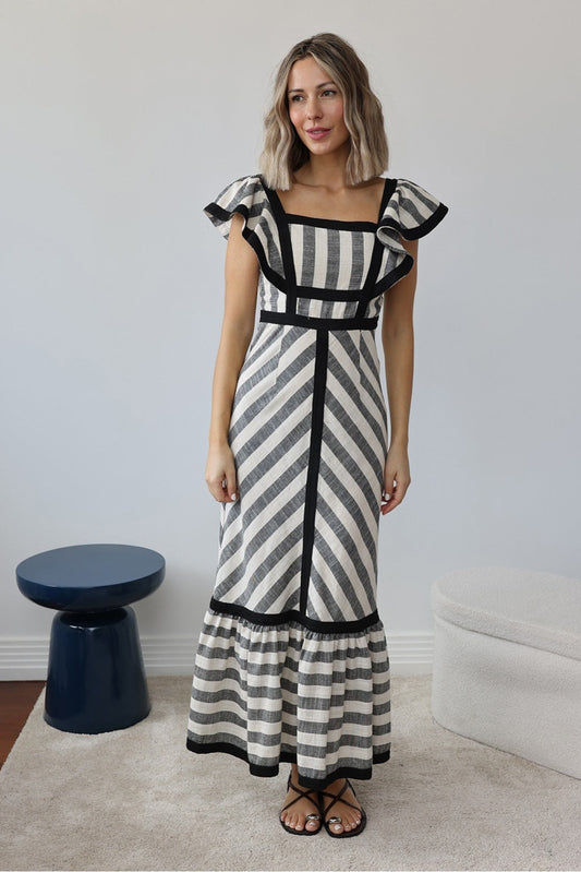 A woman wearing a structured maxi dress with a square neckline, ruffled shoulders, and a tiered hem, featuring a bold mixed-direction stripe pattern and contrast black binding.