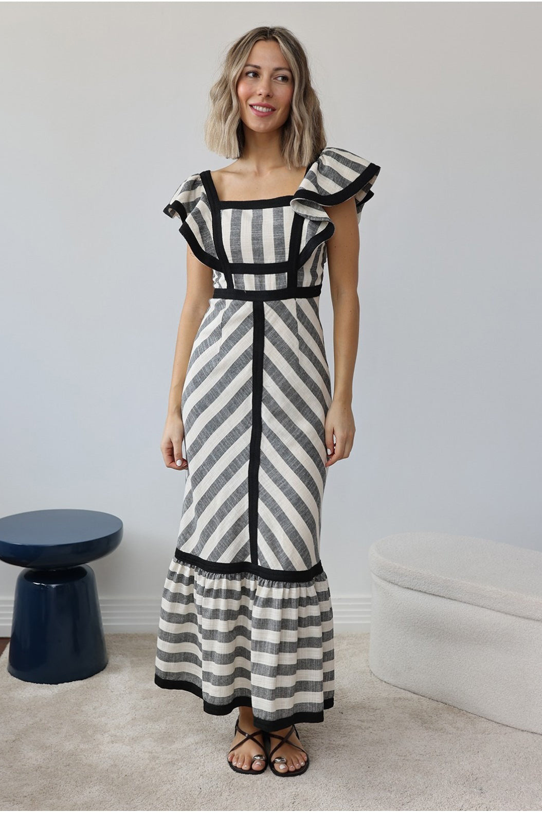 Stripe - A woman wearing a structured maxi dress with a square neckline, ruffled shoulders, and a tiered hem, featuring a bold mixed-direction stripe pattern and contrast black binding.