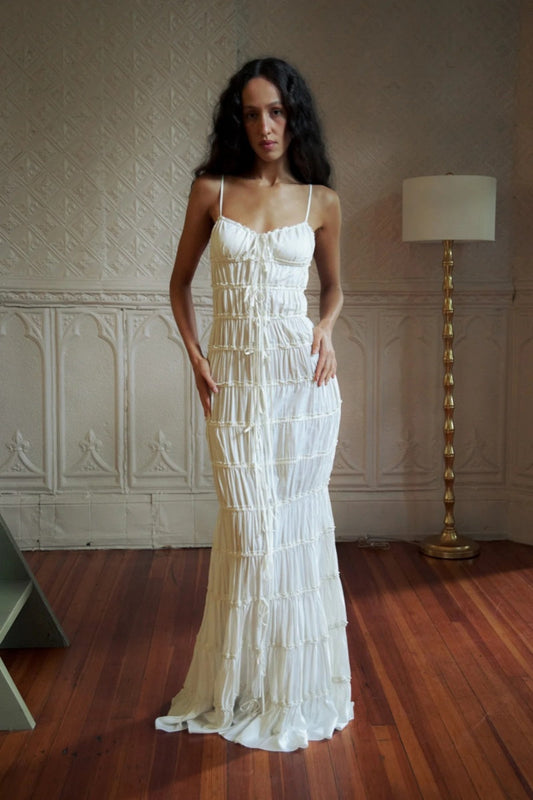 White - Woman wearing a white, floor-length maxi dress featuring all-over horizontal ruching, delicate spaghetti straps, and tie-front details down the centre.