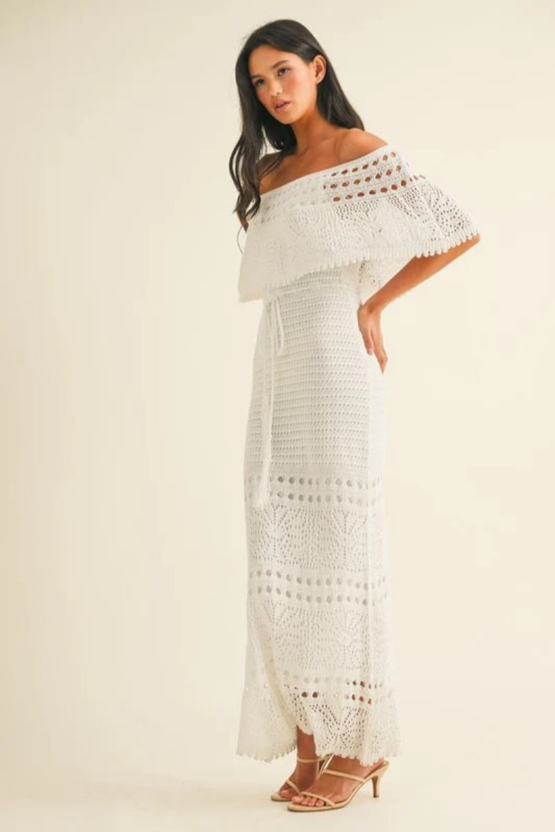White - A woman wearing a white, off-the-shoulder crochet maxi dress with a ruffle neckline and tie-waist detail, standing against a neutral studio background.