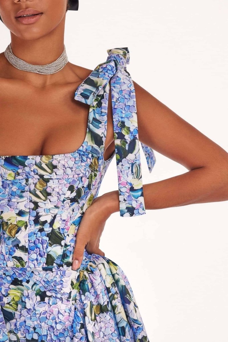 Blue - A woman models an elegant floor-length maxi dress featuring a structured corset bodice, tie-shoulder straps, and a vibrant blue and green floral print on a crisp, voluminous fabric.