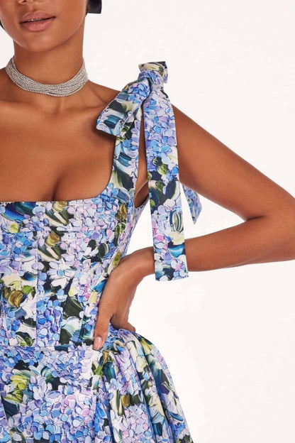 Blue - A woman models an elegant floor-length maxi dress featuring a structured corset bodice, tie-shoulder straps, and a vibrant blue and green floral print on a crisp, voluminous fabric.