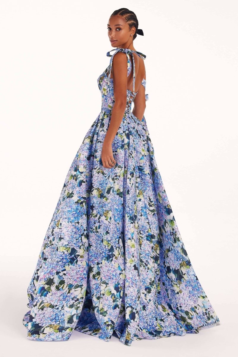 Blue - A woman models an elegant floor-length maxi dress featuring a structured corset bodice, tie-shoulder straps, and a vibrant blue and green floral print on a crisp, voluminous fabric.