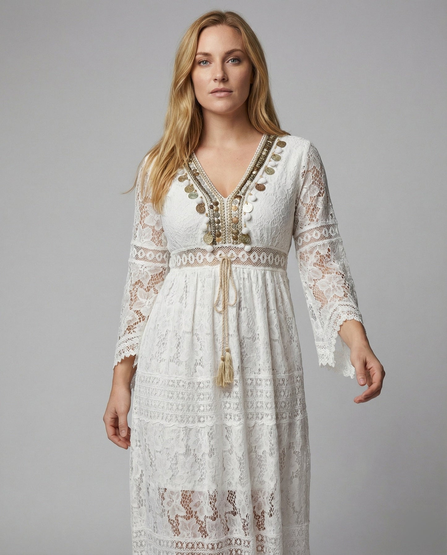 White - A woman wearing a bohemian lace maxi dress with a deep V-neckline, metallic embellishments, bell sleeves, and a tassel tie waist, holding a round woven bag.