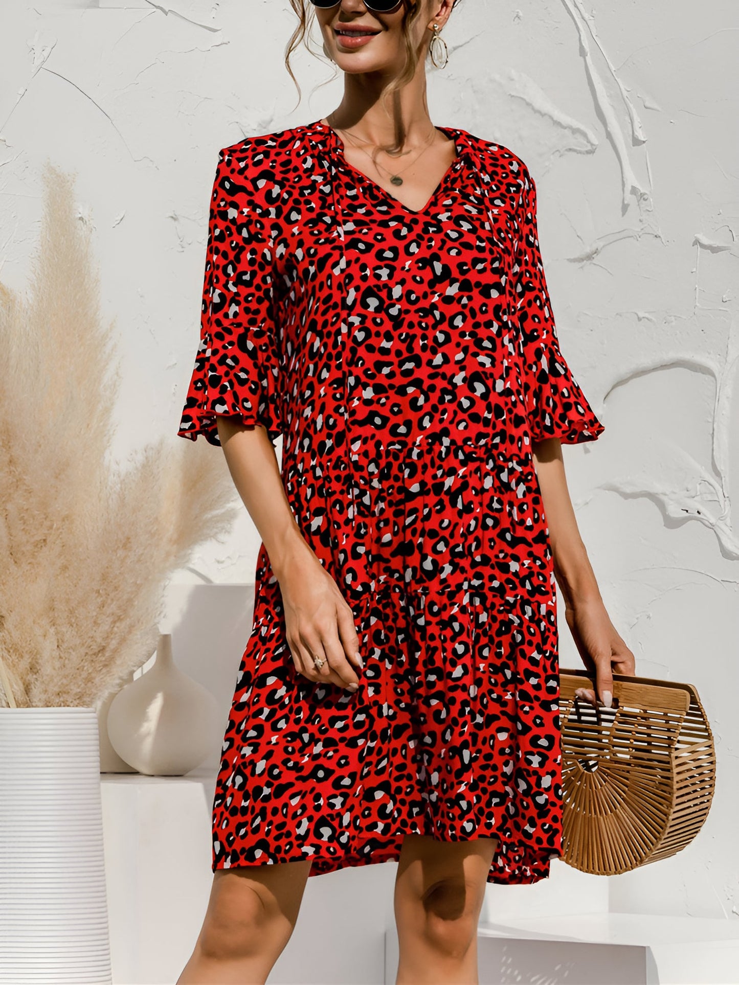 Red - Model wearing a woven leopard print v-neck tiered mini dress with flutter sleeves and neckline tie details, holding a structured bamboo handbag.
