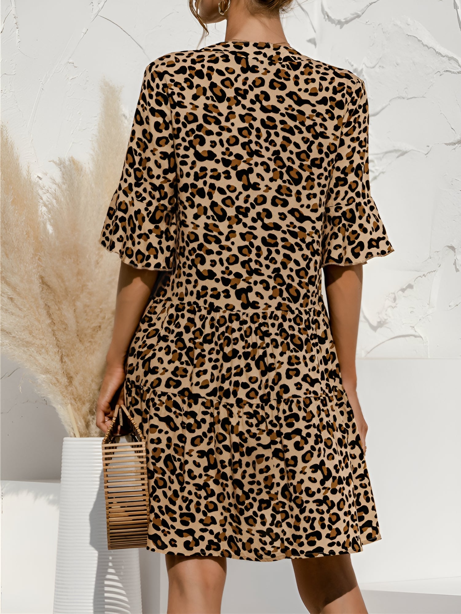 Leopard - Model wearing a woven leopard print v-neck tiered mini dress with flutter sleeves and neckline tie details, holding a structured bamboo handbag.