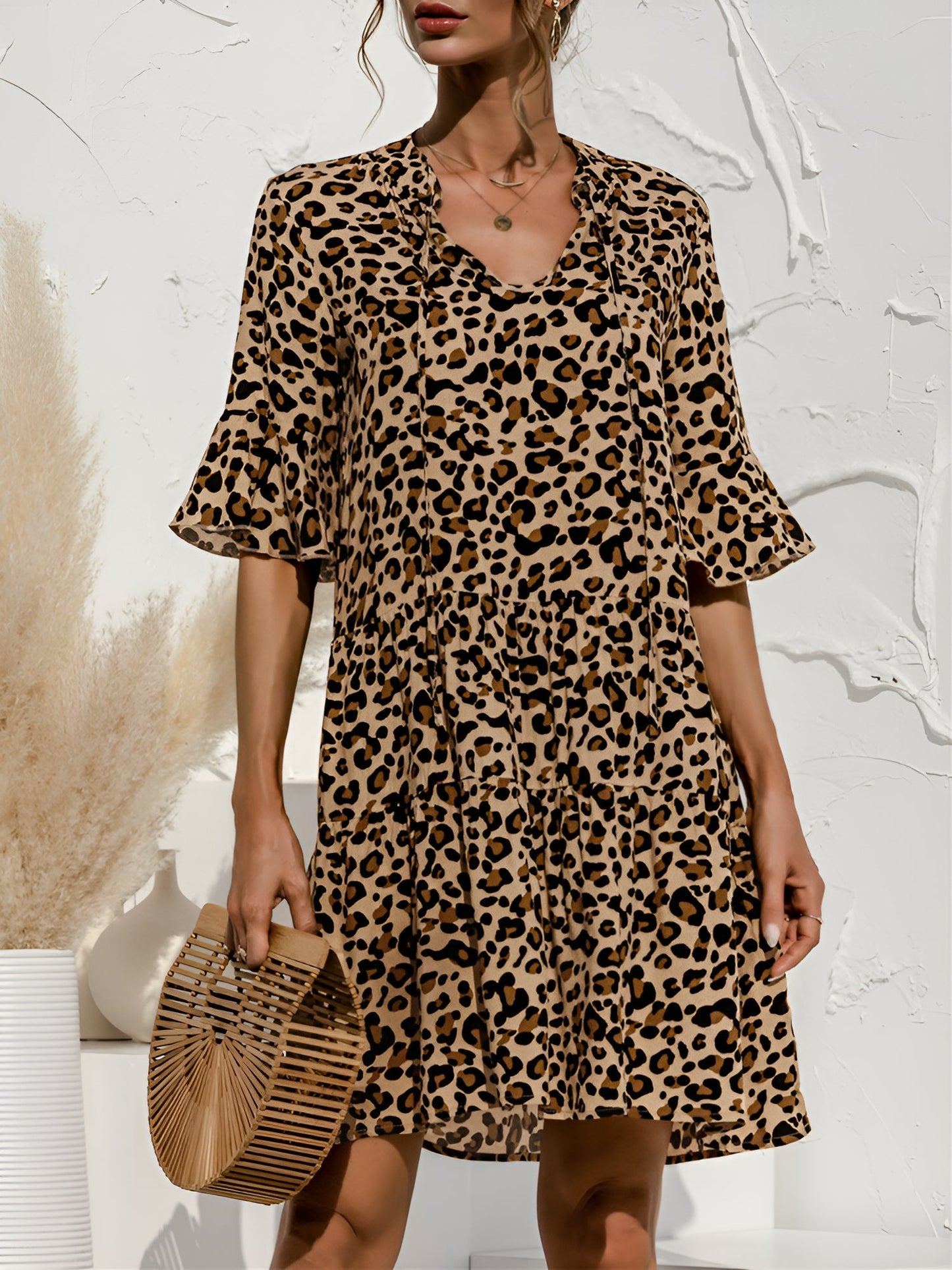 Leopard - Model wearing a woven leopard print v-neck tiered mini dress with flutter sleeves and neckline tie details, holding a structured bamboo handbag.