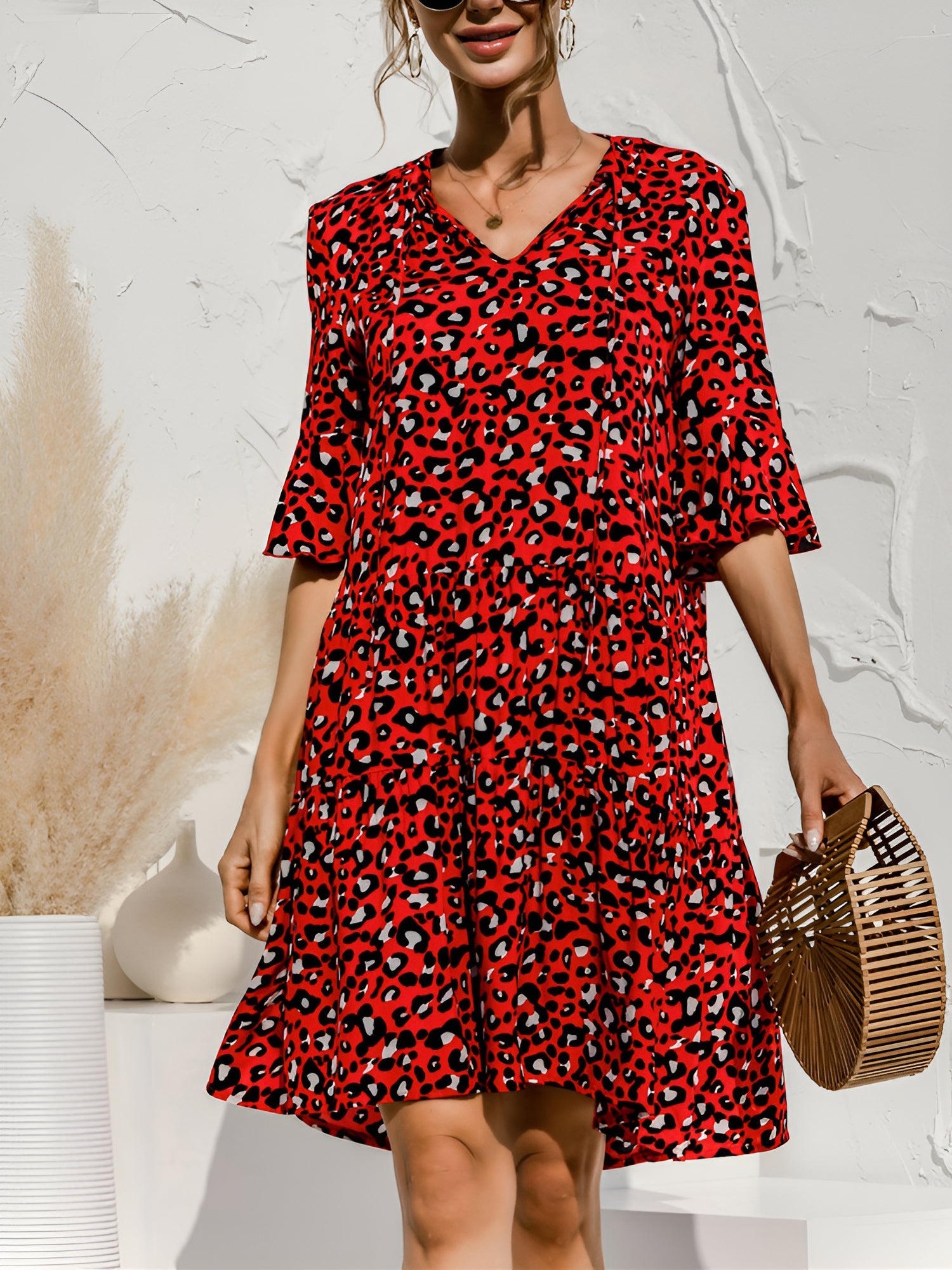 Red - Model wearing a woven leopard print v-neck tiered mini dress with flutter sleeves and neckline tie details, holding a structured bamboo handbag.
