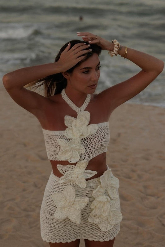 White - A woman stands on a sandy beach, wearing a form-fitting, cream crochet halter dress with a side waist cut-out and a cascade of large, three-dimensional floral appliqués.