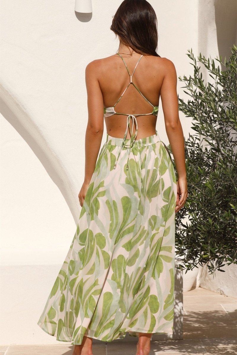 Green - A woman models a full-length halter dress with a green and cream abstract leaf print. The dress features a plunging neckline, a central ring detail at the bust, cut-outs at the waist, and a long, flowing skirt.