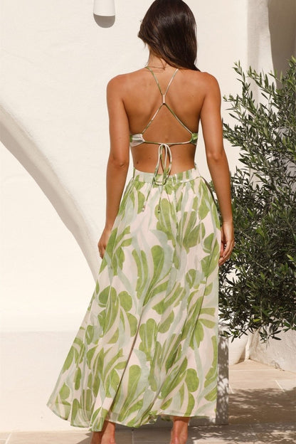 Green - A woman models a full-length halter dress with a green and cream abstract leaf print. The dress features a plunging neckline, a central ring detail at the bust, cut-outs at the waist, and a long, flowing skirt.