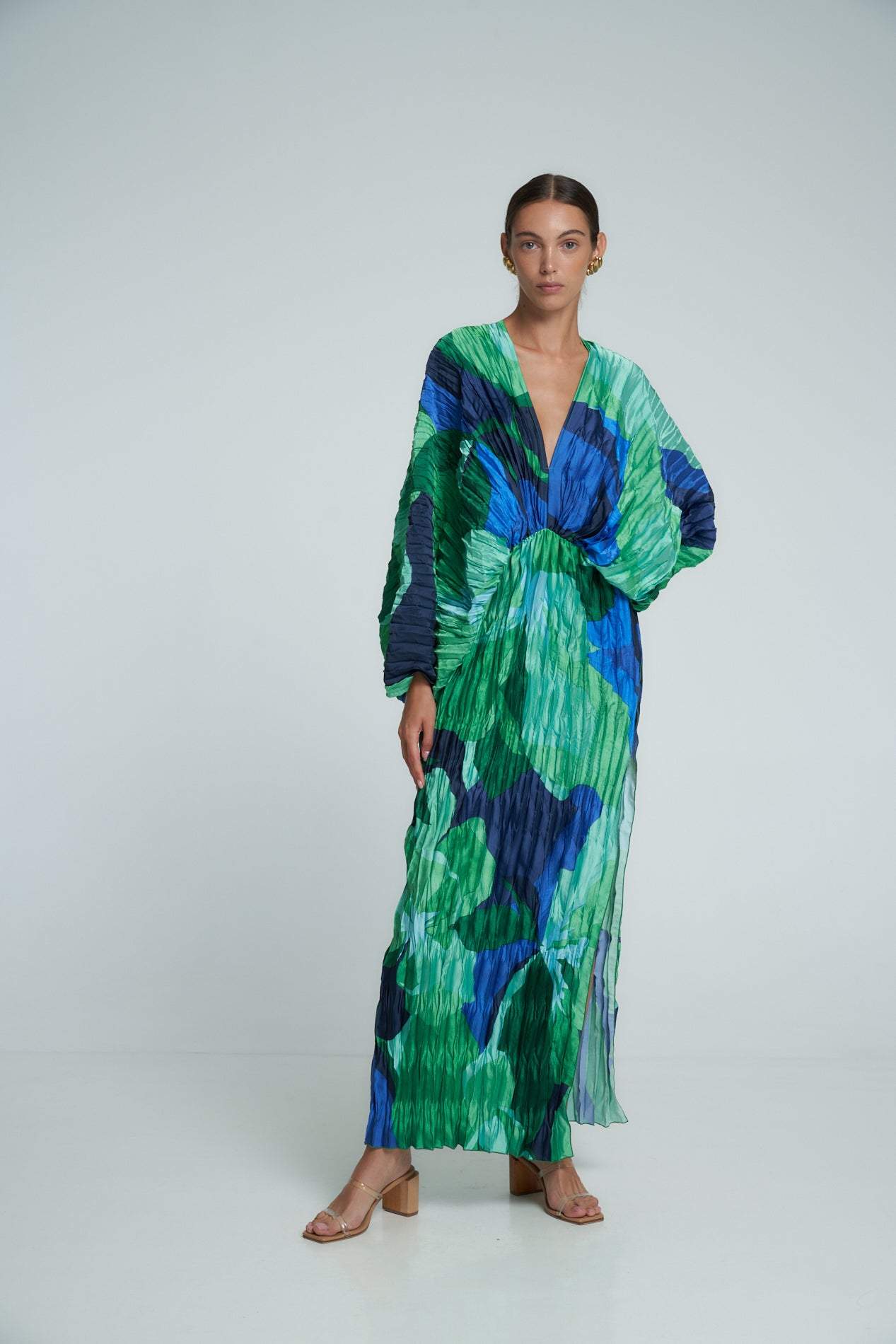 Green - A woman models a vibrant green and blue abstract print maxi dress featuring all-over plissé texture, a deep V-neckline, long voluminous sleeves, and a high side slit.
