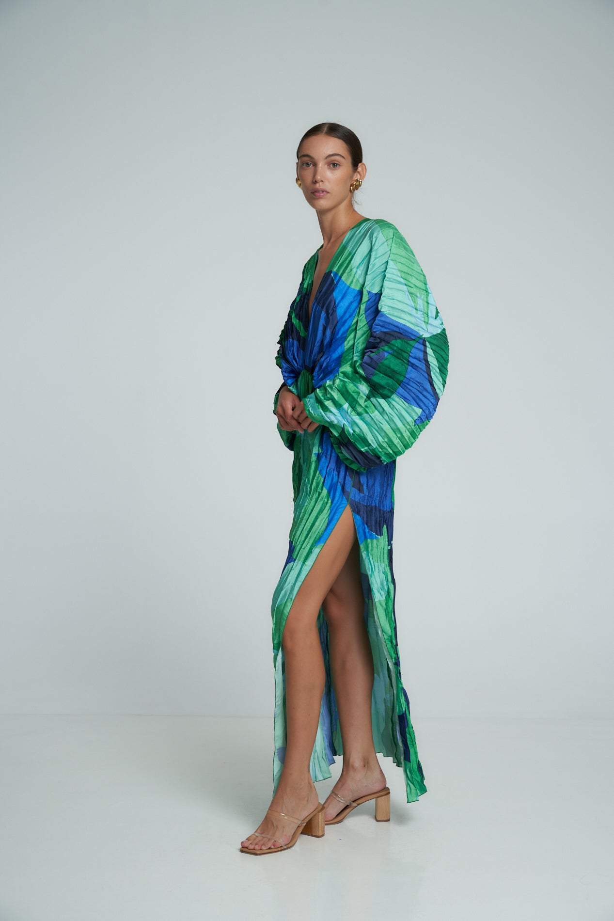 Green - A woman models a vibrant green and blue abstract print maxi dress featuring all-over plissé texture, a deep V-neckline, long voluminous sleeves, and a high side slit.