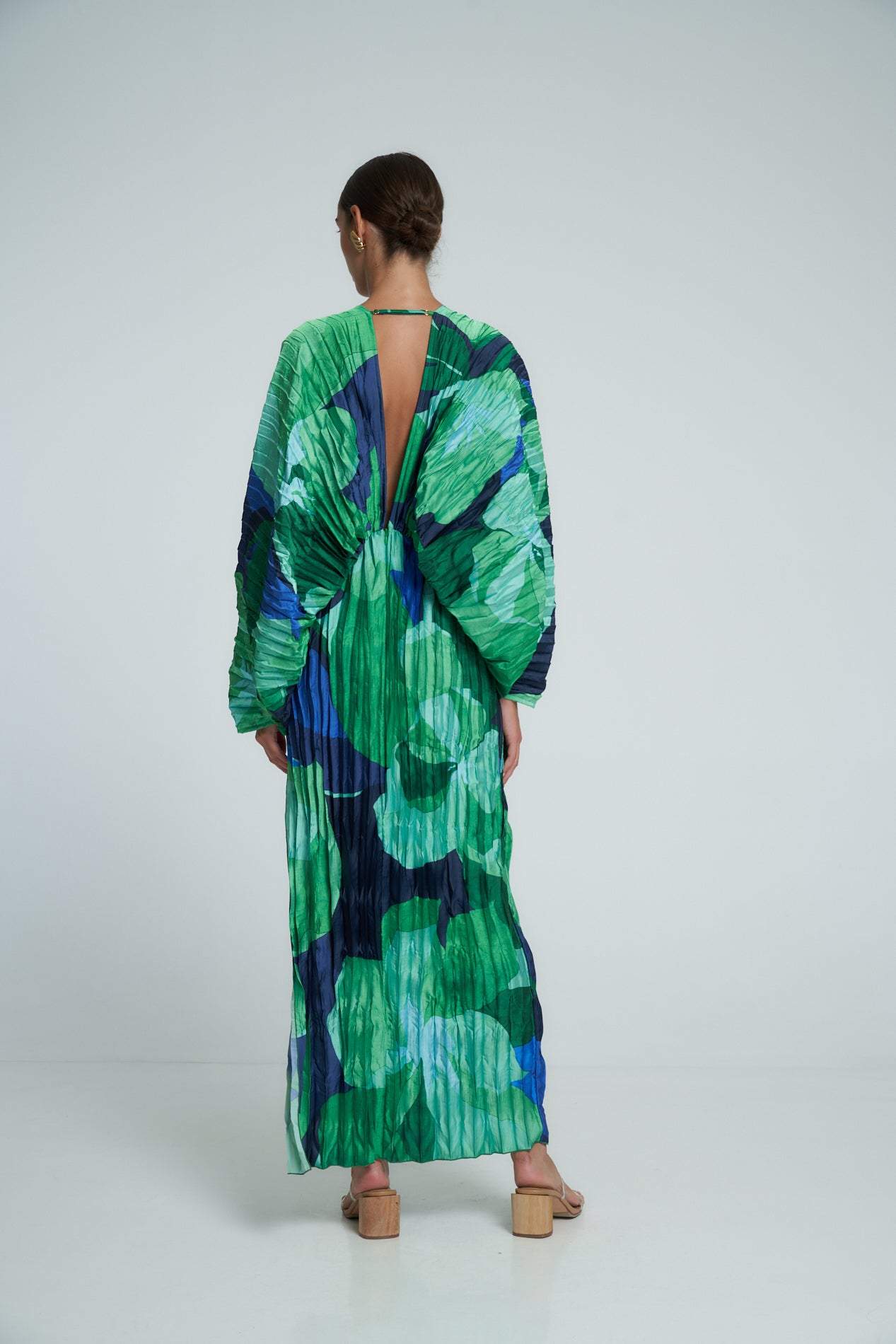 Green - A woman models a vibrant green and blue abstract print maxi dress featuring all-over plissé texture, a deep V-neckline, long voluminous sleeves, and a high side slit.