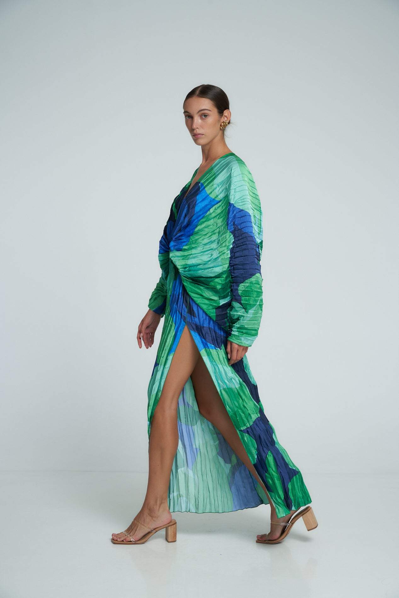 Green - A woman models a vibrant green and blue abstract print maxi dress featuring all-over plissé texture, a deep V-neckline, long voluminous sleeves, and a high side slit.