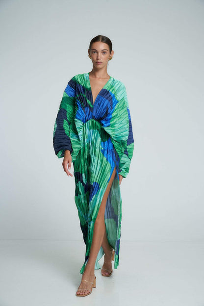 Green - A woman models a vibrant green and blue abstract print maxi dress featuring all-over plissé texture, a deep V-neckline, long voluminous sleeves, and a high side slit.
