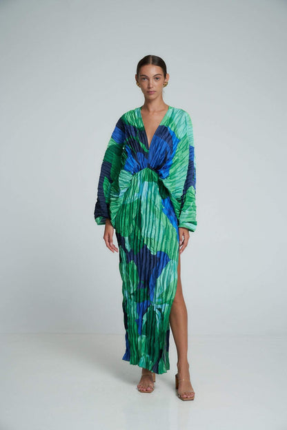 Green - A woman models a vibrant green and blue abstract print maxi dress featuring all-over plissé texture, a deep V-neckline, long voluminous sleeves, and a high side slit.
