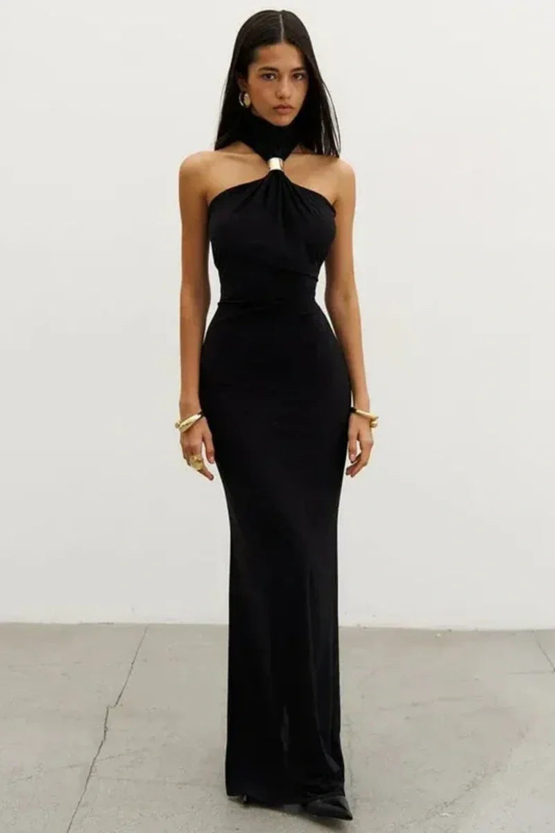 Black - Woman wearing a white form-fitting halterneck maxi dress with a gold ring detail at the neckline, standing indoors.