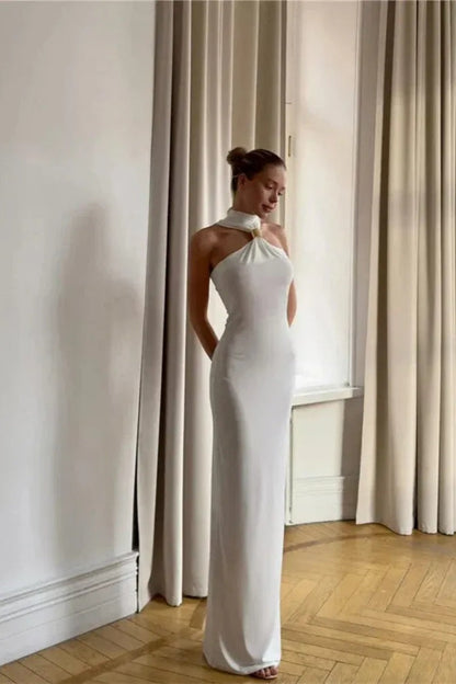 White - Woman wearing a white form-fitting halterneck maxi dress with a gold ring detail at the neckline, standing indoors.