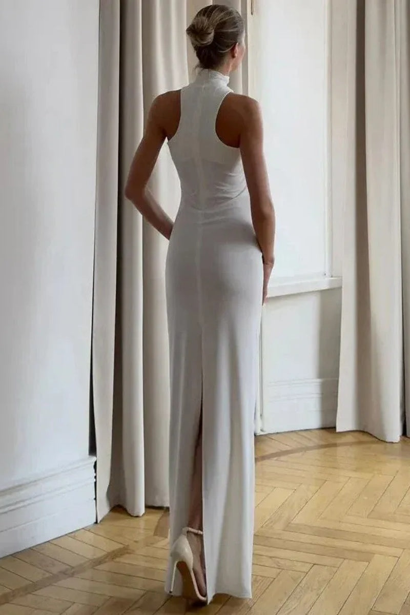 White - Woman wearing a white form-fitting halterneck maxi dress with a gold ring detail at the neckline, standing indoors.