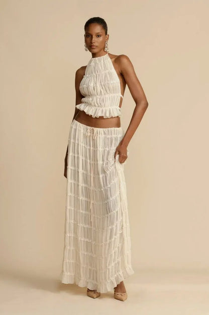 White - A model wearing a white, two-piece matching set featuring a cropped halter top and a flowing maxi skirt, both designed with a semi-sheer, crinkled fabric and horizontal ruched tiers.