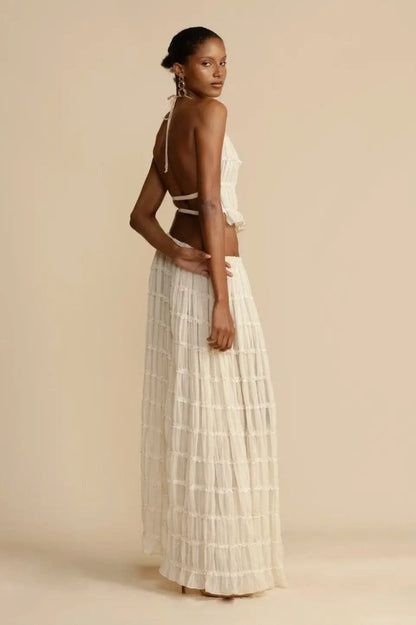White - A model wearing a white, two-piece matching set featuring a cropped halter top and a flowing maxi skirt, both designed with a semi-sheer, crinkled fabric and horizontal ruched tiers.