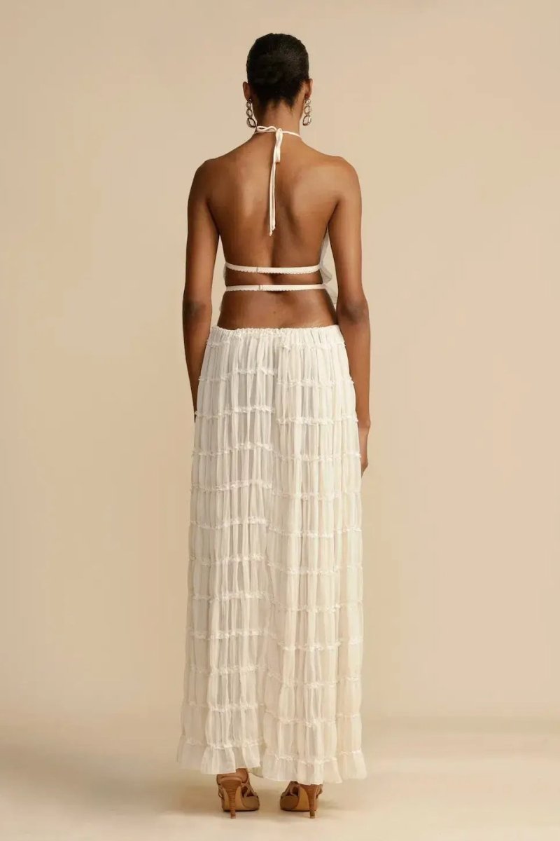 White - A model wearing a white, two-piece matching set featuring a cropped halter top and a flowing maxi skirt, both designed with a semi-sheer, crinkled fabric and horizontal ruched tiers.