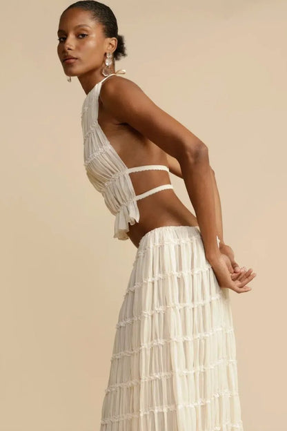 White - A model wearing a white, two-piece matching set featuring a cropped halter top and a flowing maxi skirt, both designed with a semi-sheer, crinkled fabric and horizontal ruched tiers.