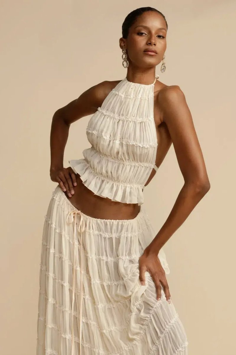 White - A model wearing a white, two-piece matching set featuring a cropped halter top and a flowing maxi skirt, both designed with a semi-sheer, crinkled fabric and horizontal ruched tiers.