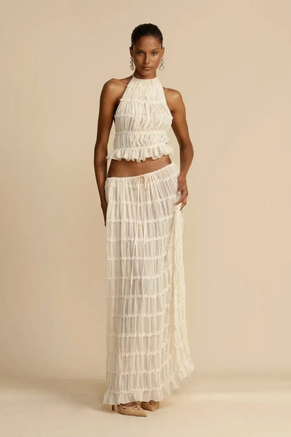 White - A model wearing a white, two-piece matching set featuring a cropped halter top and a flowing maxi skirt, both designed with a semi-sheer, crinkled fabric and horizontal ruched tiers.