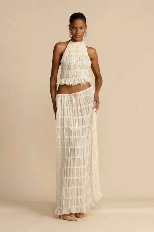 White - A model wearing a white, two-piece matching set featuring a cropped halter top and a flowing maxi skirt, both designed with a semi-sheer, crinkled fabric and horizontal ruched tiers.