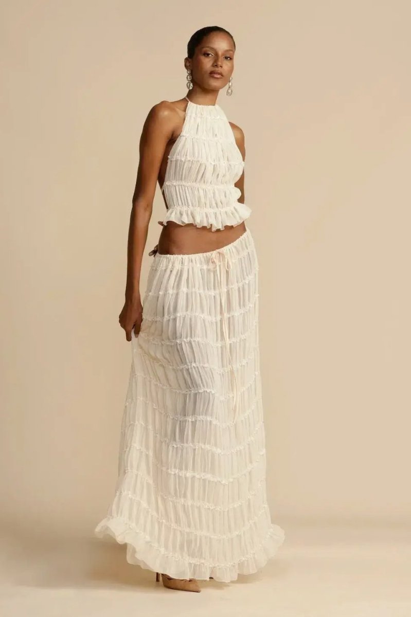 White - A model wearing a white, two-piece matching set featuring a cropped halter top and a flowing maxi skirt, both designed with a semi-sheer, crinkled fabric and horizontal ruched tiers.