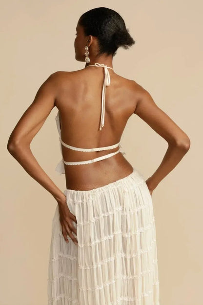 White - A model wearing a white, two-piece matching set featuring a cropped halter top and a flowing maxi skirt, both designed with a semi-sheer, crinkled fabric and horizontal ruched tiers.
