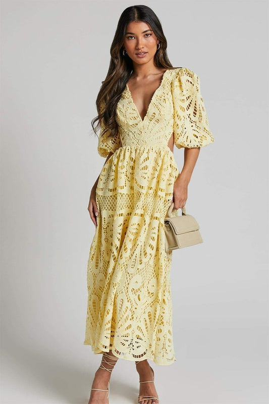 Yellow - A model wearing a pale yellow, maxi-length dress featuring an all-over intricate cut-out lace pattern, a deep V-neckline, short puff sleeves, and side waist cut-outs, accessorised with strappy heels and a small handbag.